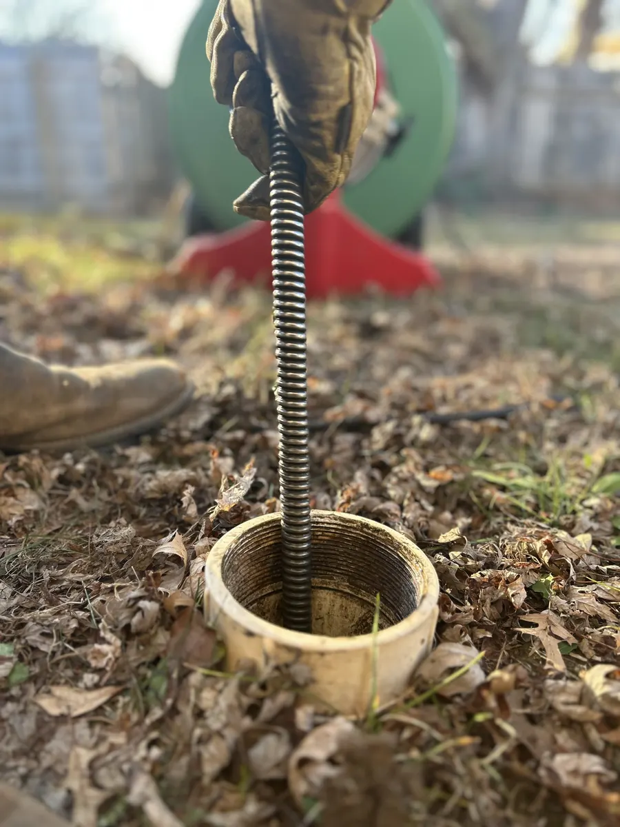 A Mosley technician’s gloved hand feeding a drain auger cable into a sewer cleanout in a leaf-covered yard